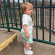 Kasen is registered to the contest to win money with this photo: child, toddler, outdoor, fence, clothing, shorts, tshirt, sneakers, concrete, side_view, green, metal, bare_trees, sky, rocks, serious_expression, person, standing, daytime, urban