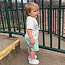 child, toddler, outdoor, fence, clothing, shorts, tshirt, sneakers, concrete, side_view, green, metal, bare_trees, sky, rocks, serious_expression, person, standing, daytime, urban