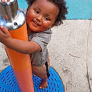 Zyair joined the competition — help win amazing prizes! barefoot, blue_surface, child, clothing, concrete, curly_hair, fun, gray_surface, happy, holding, metal, orange, outdoor, person, play, playground, pole, smiling, toddler, young_child