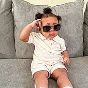Prince is registered to the contest to win money with this photo: toddler, child, sunglasses, white_clothing, sneakers, socks, couch, gray_couch, indoor, hairstyle, curly_hair, pose, cute, fashion, casual, seat, young_child, person, portrait, relaxed