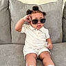 toddler, child, sunglasses, white_clothing, sneakers, socks, couch, gray_couch, indoor, hairstyle, curly_hair, pose, cute, fashion, casual, seat, young_child, person, portrait, relaxed