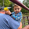 child, toddler, adult, piggyback, outdoor, playground, zoo, enclosure, toy_drill, yellow_toy, blue_pajamas, forest, trees, railings, family, portrait, serious_expression, sitting, daylight, recreation
