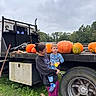 Declan is registered to the contest to win money with this photo: child, toddler, adult, person, pumpkins, pumpkin_festival, truck, flatbed_truck, vehicle, tire, outdoor, autumn, fall, farm, trees, cloudy_sky, grass, portrait, holding_child, colorful_clothing