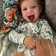 Declan joined the competition — help win amazing prizes! baby, child, infant, smiling, laughing, portrait, face, eyes, mouth, blanket, pajamas, sleepwear, patterned_clothing, lying_down, indoor, cozy, couch, adult_hand, playful, close_up
