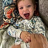 baby, child, infant, smiling, laughing, portrait, face, eyes, mouth, blanket, pajamas, sleepwear, patterned_clothing, lying_down, indoor, cozy, couch, adult_hand, playful, close_up