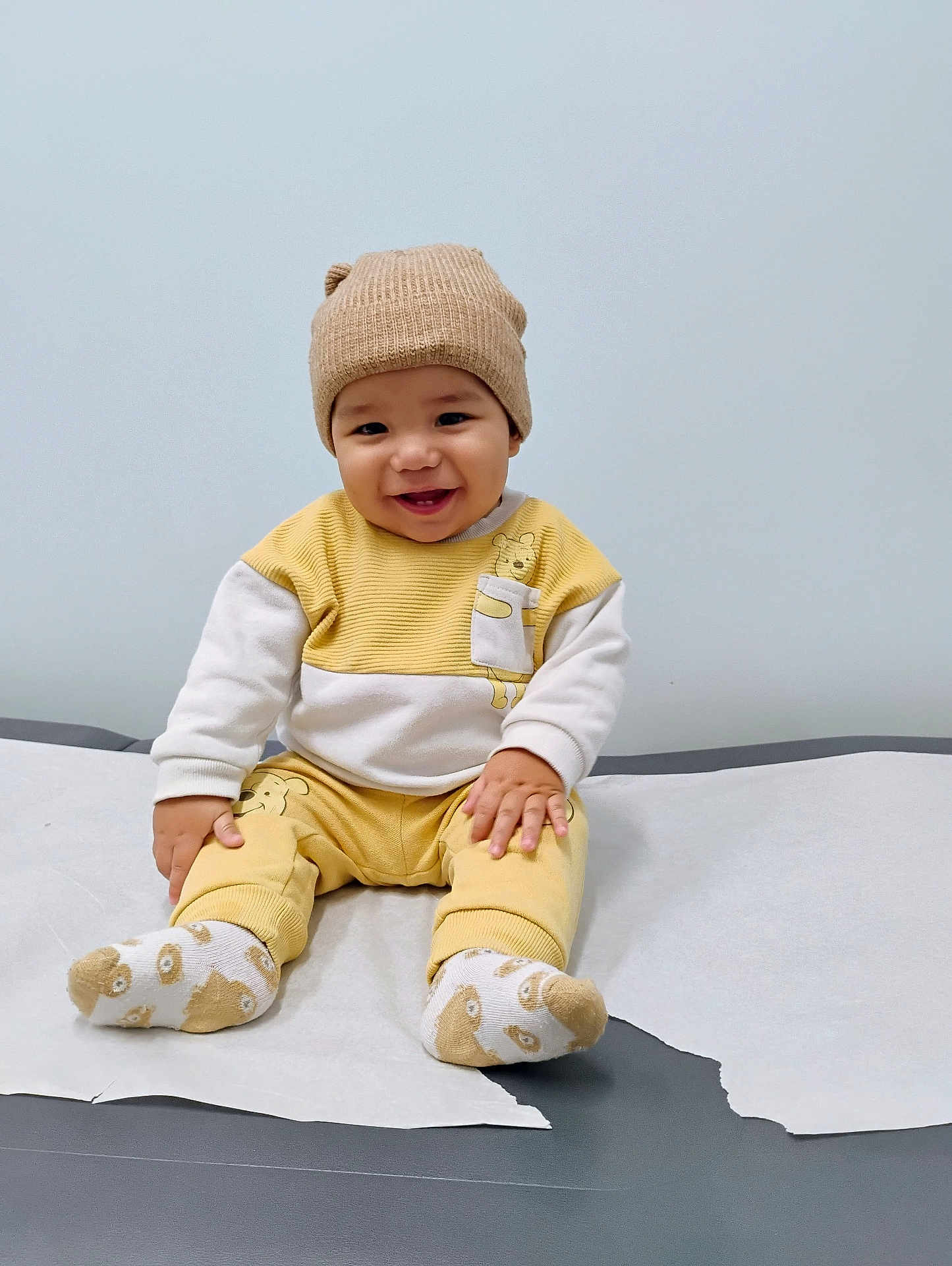 Dylan is registered to the contest to win money with this photo: baby, beige, child, clothing, cute, face, happy, hat, indoors, infant, medical_table, paper_cover, portrait, sitting, smiling, socks, warm_clothes, white, yellow, young_child