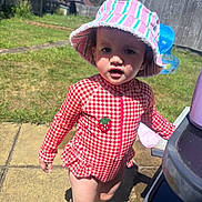 Wren is registered to the contest to win money with this photo: toddler, child, swimsuit, sun_hat, red_gingham, strawberry_patch, outdoor, backyard, grass, pavement, plastic_toy, sunny, daylight, curious, barefoot, person, portrait, summer, play, garden_fence