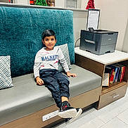 Thejus joined the competition — help win amazing prizes! child, boy, sitting, bench, cushion, printer, books, indoor, casual_clothing, holiday_decorations, tinsel, floor_tiles, modern_furniture, curious_expression, black_pants, white_shirt, sneakers, side_table, room, portrait