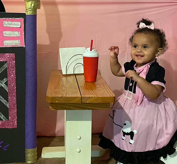 Khalani is registered to the contest to win money with this photo: art, baby, baby_toddler_clothing, child, desk, drawer, event, happy, hat, joy, magenta, office_supplies, person, room, sitting, sleeve, smile, standing, table, tableware