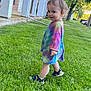 Uhtred is registered to the contest to win money with this photo: toddler, child, grass, outdoor, building, sneakers, tie_dye, smile, walking, greenery, pillars, brick_wall, cheeky, curly_hair, daylight, side_view, casual_clothing, person, young_child, playful