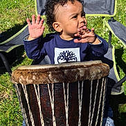 Qassim a rejoint le concours — aidez-le/la à gagner de superbes lots ! blue_clothing, camping_chair, child, curly_hair, daylight, drum, expression, grass, hands, musical_instrument, nature, outdoor, parked_cars, person, playing, seat, shoes, sunlight, toddler, tree