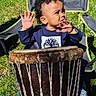 blue_clothing, camping_chair, child, curly_hair, daylight, drum, expression, grass, hands, musical_instrument, nature, outdoor, parked_cars, person, playing, seat, shoes, sunlight, toddler, tree