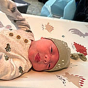 Raymond is registered to the contest to win money with this photo: newborn, baby, infant, hat, clothing, dinosaur_pattern, mattress, cute, lying_down, tongue_out, soft_light, indoors, bedroom, knitted_hat, onesie, sleepwear, child, small, adorable, portrait