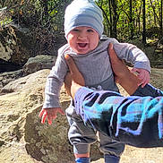 Michael is registered to the contest to win money with this photo: baby, child, outdoor, nature, rock, tree, greenery, sunlight, hat, clothing, socks, happy, smile, person, hands, forest, daylight, cute, holding, playful