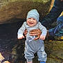 Michael joined the competition — help win amazing prizes! baby, smiling, child, adult, hat, cap, nature, rock, stream, water, outdoor, clothing, blue_socks, smile, happy, person, holding, supporting, forest, sunlight