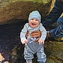 baby, child, smiling, adult, hand, rock, water, creek, outdoor, nature, clothing, beanie, blue_socks, gray_pants, plaid_shirt, cap, tattoo, sunlight, happy, support