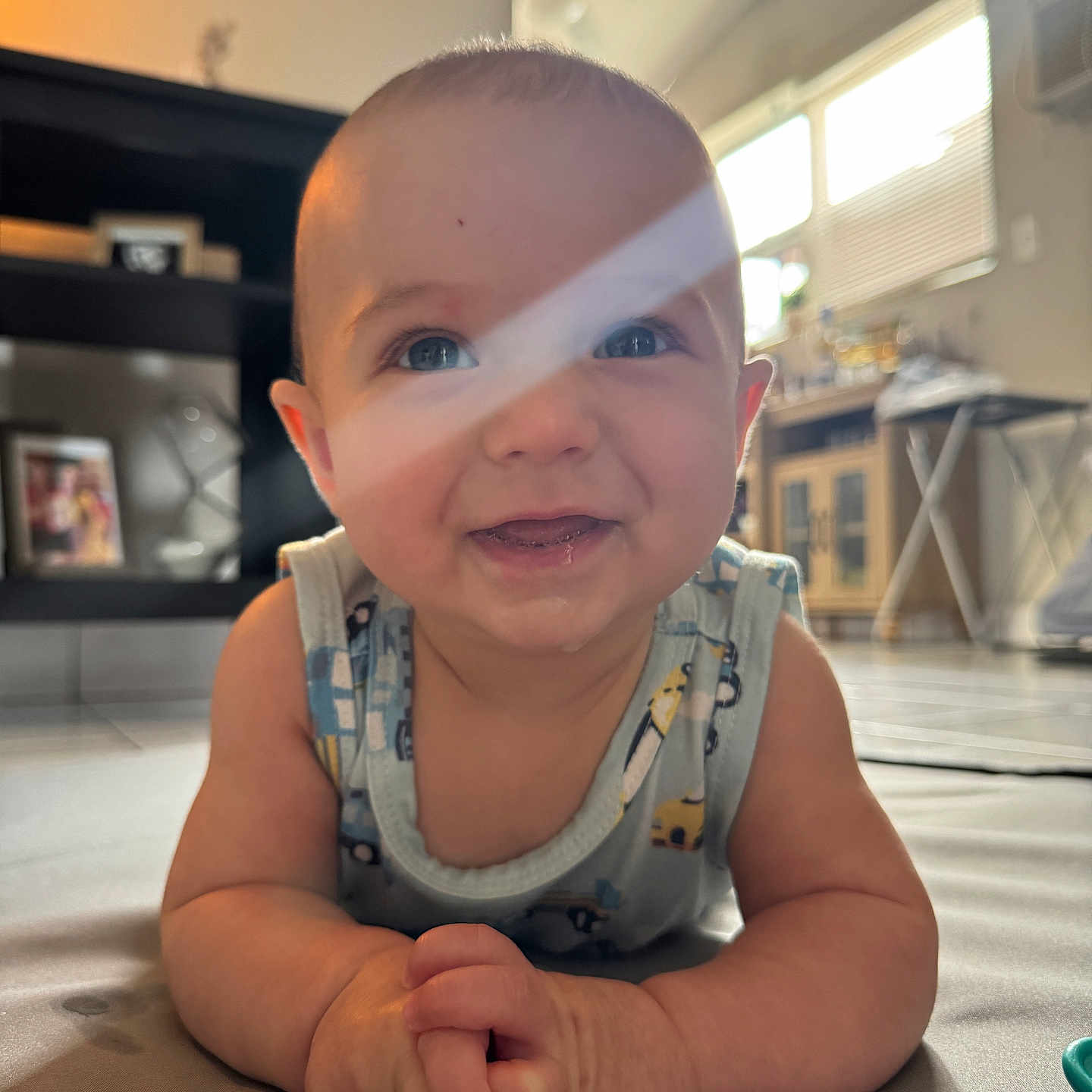 Darell joined the competition — help win amazing prizes! baby, infant, child, smiling, portrait, indoor, living_room, play_mat, sitting, crawling, onesie, blue_eyes, face, hands, arms, sunlight, window, shelves, home, happy