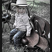 Lainey is registered to the contest to win money with this photo: child, toddler, cowboy_hat, striped_shirt, belt, jeans, saddle, grass, wagon_wheel, outdoor, western, portrait, serious_expression, hat, footwear, person, nature, vintage, rural, rustic