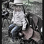 child, toddler, cowboy_hat, striped_shirt, belt, jeans, saddle, grass, wagon_wheel, outdoor, western, portrait, serious_expression, hat, footwear, person, nature, vintage, rural, rustic