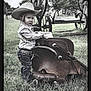 child, cowboy_hat, striped_shirt, jeans, belt, saddle, grass, outdoor, fence, wheel, tree, bench, person, portrait, western, ranch, daylight, young, cowboy, rural