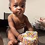 baby, child, cake, sprinkles, frosting, table, food, diaper, hand, face, curly_hair, indoor, celebration, messy, cute, person, sitting, birthday, sweet, dessert