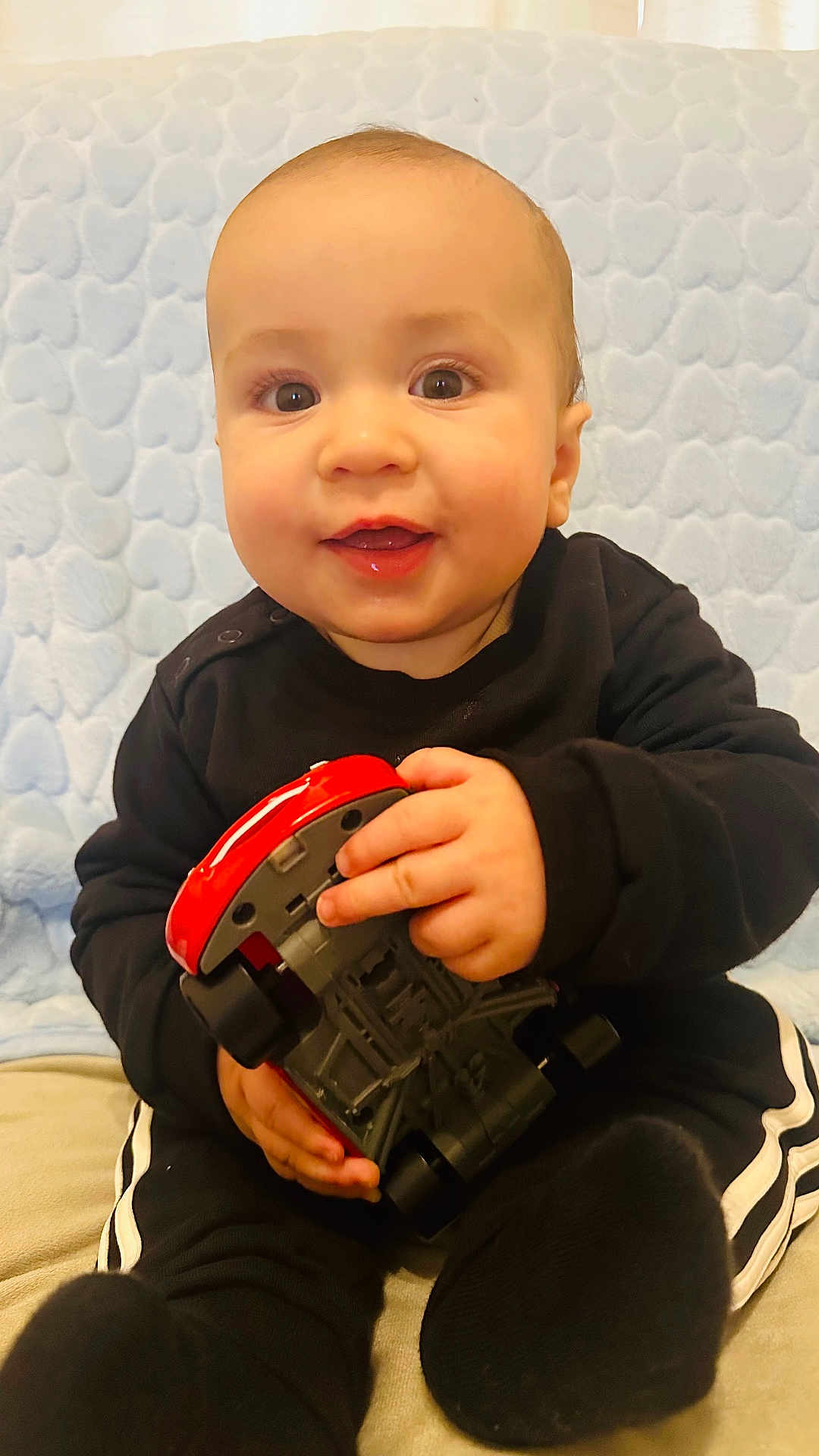 Enzo joined the competition — help win amazing prizes! baby, child, toy, car, black_clothing, smiling, sitting, indoors, blanket, heart_pattern, cute, happy, portrait, person, infant, playful, hands, feet, face, closeup