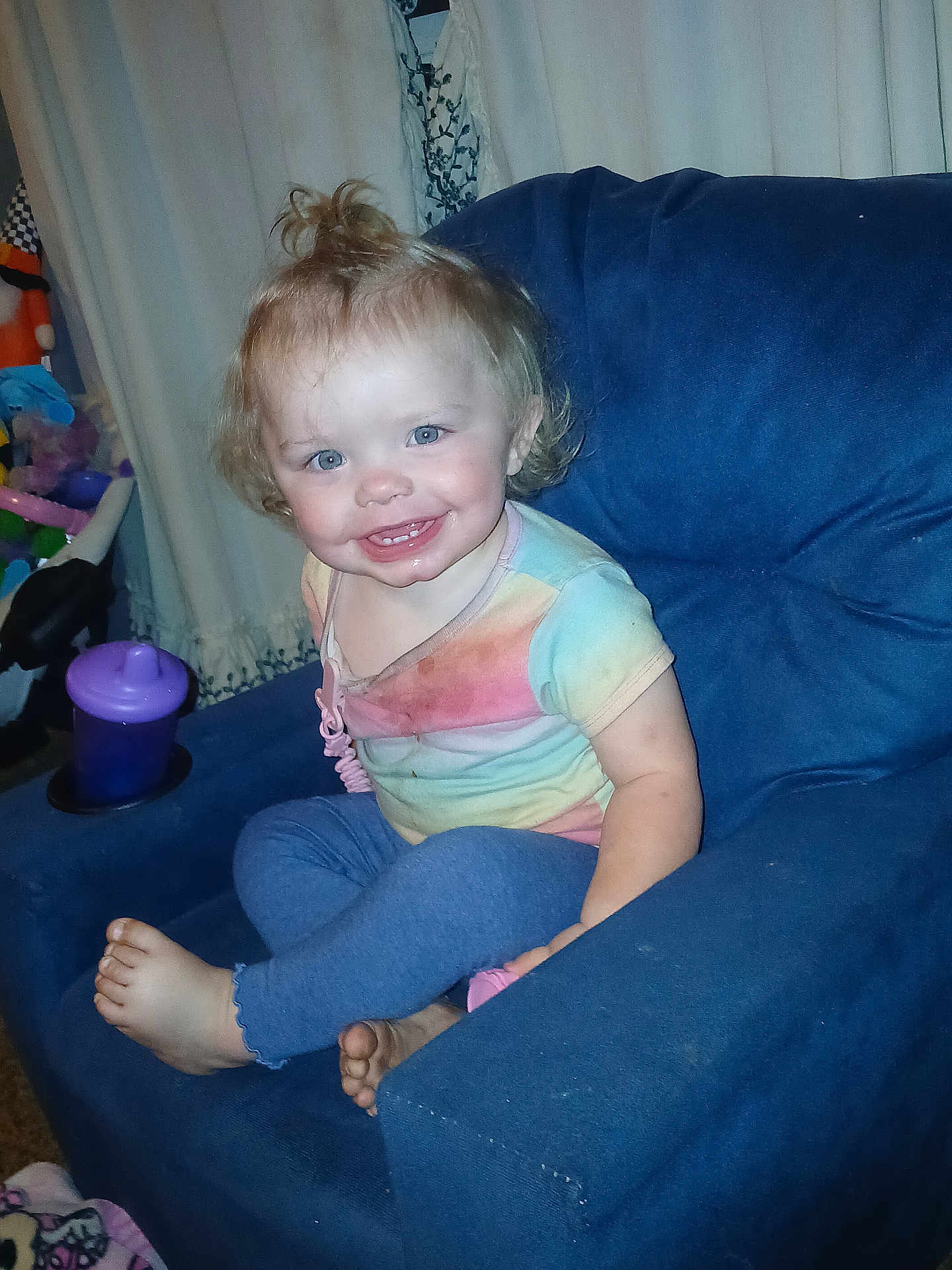 Stormie is registered to the contest to win money with this photo: toddler, child, smile, blue_eyes, rainbow_shirt, leggings, barefoot, armchair, indoor, curtains, sippy_cup, toys, happy, hair, person, seated, candid, home, furniture, playroom