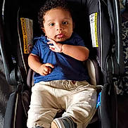Hasani is registered to the contest to win money with this photo: baby, infant, child, car_seat, carrier, seat, blue_shirt, beige_pants, sneakers, curly_hair, face, eyes, hands, portrait, indoor, blanket, warning_label, fabric, footwear, person