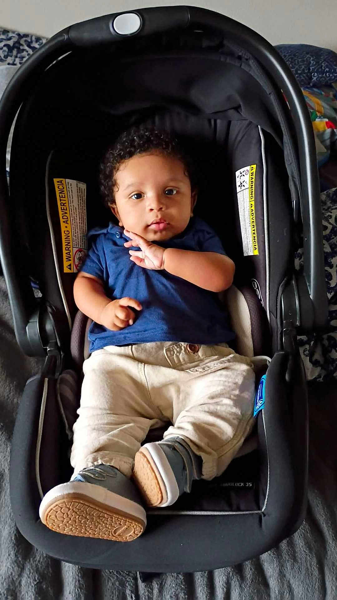 Hasani is registered to the contest to win money with this photo: baby, infant, child, car_seat, carrier, seat, blue_shirt, beige_pants, sneakers, curly_hair, face, eyes, hands, portrait, indoor, blanket, warning_label, fabric, footwear, person