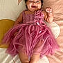 baby, child, smiling, dress, pink, headband, flower, curly_hair, lying_down, happy, cute, infant, tulle, fabric, barefoot, skin, indoors, portrait, person, bedspread