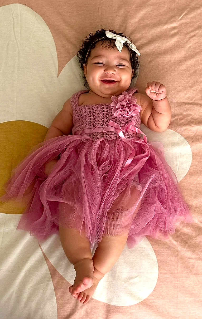 Olivia is registered to the contest to win money with this photo: baby, child, smiling, dress, pink, headband, flower, curly_hair, lying_down, happy, cute, infant, tulle, fabric, barefoot, skin, indoors, portrait, person, bedspread