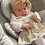 baby, infant, headband, skirt, white_blouse, cushion, plush, soft_fabric, child, portrait, indoors, cute, serious_expression, sitting, hand, face, clothing, patterned_skirt, comfort, headwear