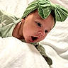 baby, infant, headband, green_bow, curious, wide_eyes, mouth_open, blanket, white_blanket, closeup, face, skin, cute, soft_texture, lying_down, indoors, child, adorable, expression, young