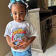 Noelle is registered to the contest to win money with this photo: toddler, child, indoor, person, clothing, tshirt, shorts, headband, hair, standing, floor, cabinet, television, wooden_furniture, expression, hands, portrait, casual_wear, cute, young_child