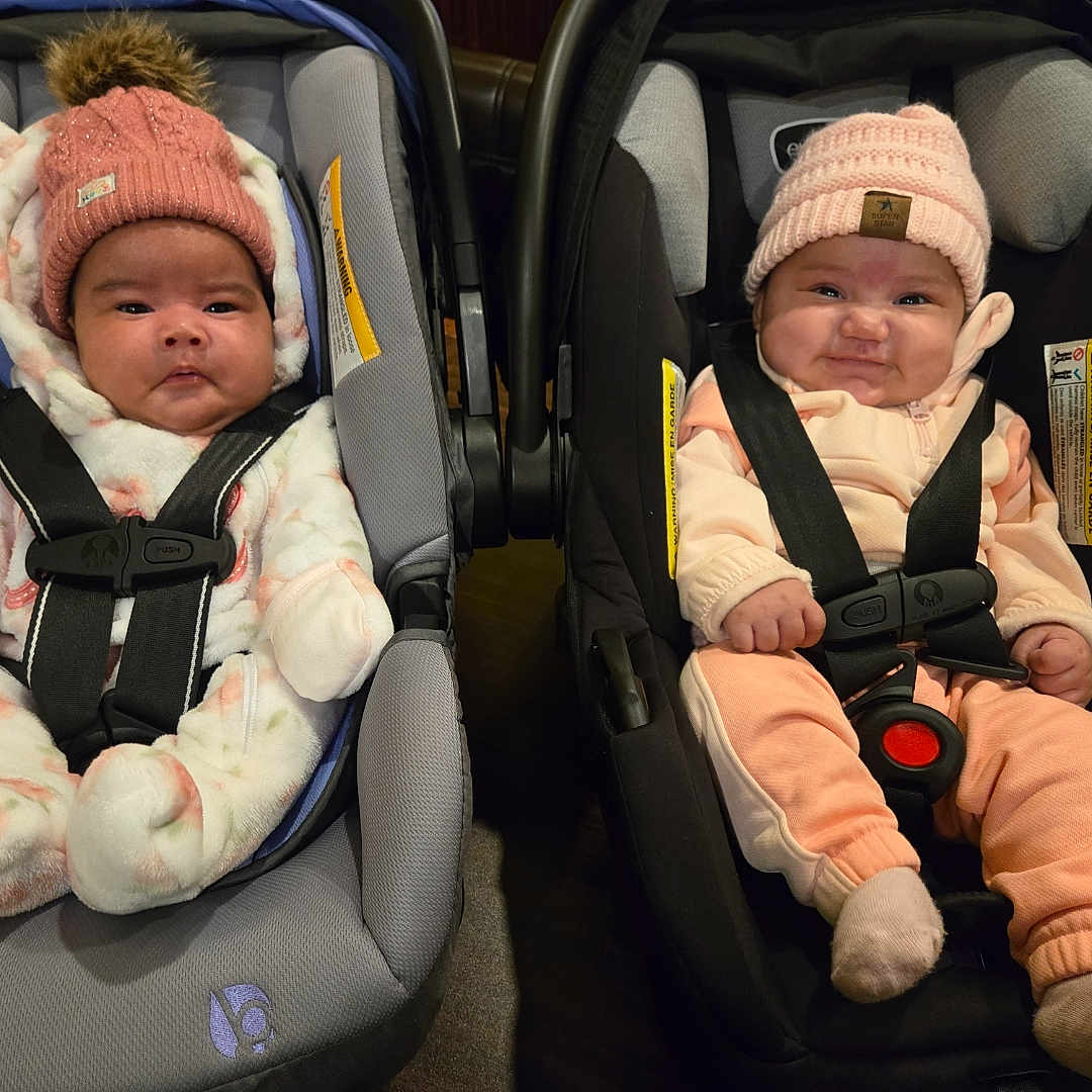 Selaima is registered to the contest to win money with this photo: adorable, baby, car_seat, child, cozy, cute, indoor, infant, knit_hat, person, pink_hat, portrait, seat_belt, serious_face, sitting, smiling, straps, two_babies, warm_clothing, winter_clothing