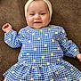 baby, child, smiling, headband, bow, blue_dress, checkered_pattern, flowers, lying_down, brown_background, cute, infant, happy, clothing, portrait, young_child, indoors, person, head, face