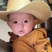 Stetson is registered to the contest to win money with this photo: baby, child, cowboy_hat, hoodie, person, indoor, kitchen, curious, wide_eyes, holding, face, apparel, clothing, headwear, portrait, infant, human, cute, expression, casual
