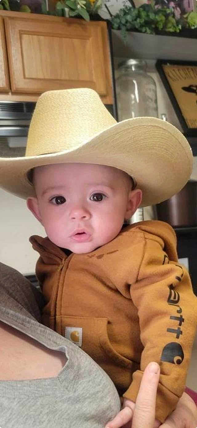 Stetson is registered to the contest to win money with this photo: baby, child, cowboy_hat, hoodie, person, indoor, kitchen, curious, wide_eyes, holding, face, apparel, clothing, headwear, portrait, infant, human, cute, expression, casual