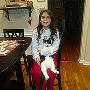Gracie joined the competition — help win amazing prizes! girl, cat, chair, table, phone, wooden_floor, indoor, smiling, long_hair, red_pants, gray_hoodie, black_and_white_cat, framed_photos, wall_art, holiday_decor, table_runner, sitting, happy, home, cozy