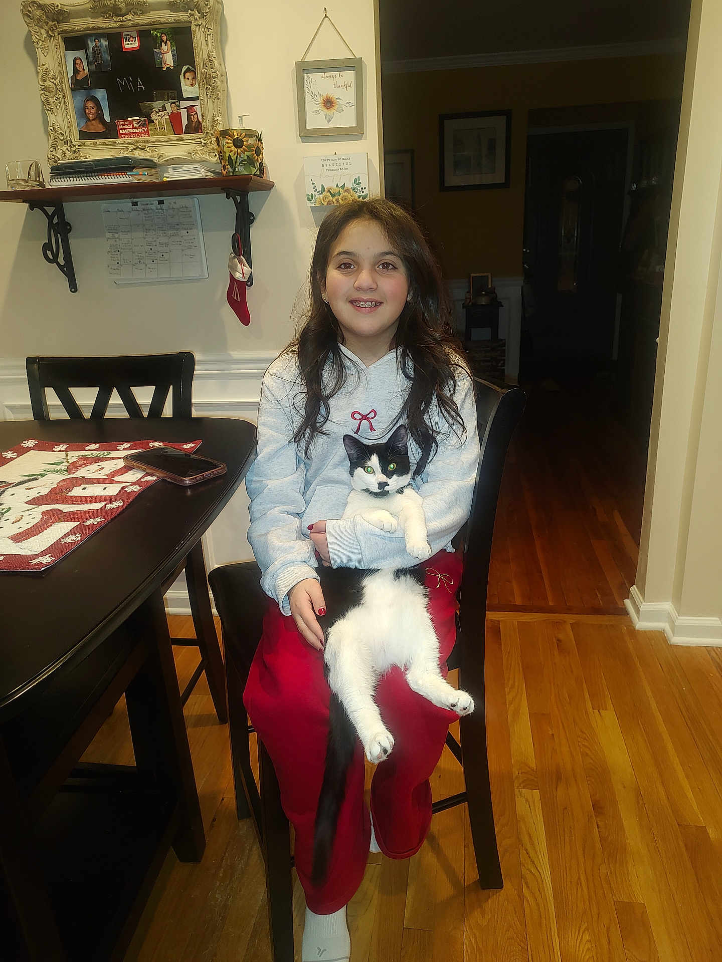 Gracie joined the competition — help win amazing prizes! girl, cat, chair, table, phone, wooden_floor, indoor, smiling, long_hair, red_pants, gray_hoodie, black_and_white_cat, framed_photos, wall_art, holiday_decor, table_runner, sitting, happy, home, cozy