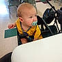 baby, pacifier, high_chair, yellow_shirt, indoor, table, chair, floor, child, infant, person, seated, looking_away, daylight, curious, plastic, feeding, toddler, casual, room