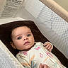baby, infant, crib, onesie, mesh, bedding, patterned_clothing, child, lying_down, indoor, portrait, cute, small_hands, wide_eyes, curious_expression, soft_lighting, head, face, newborn, sleeping_area