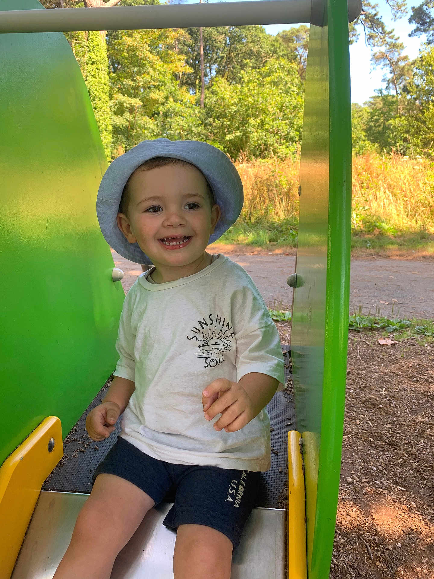 Gabin participe au concours pour gagner de l'argent avec cette photo : child, toddler, smiling, slide, playground, hat, shorts, outdoor, green, sunlight, trees, nature, summer, happy, park, playing, clothing, fun, person, recreation