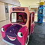 toddler, child, kiddie_ride, toy_car, mall, indoor, pink, purple, smiling, play, entertainment, floor, ceiling, lights, window, person, face, bow, clothing, fun