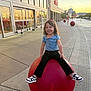 child, smiling, outdoor, red_sphere, sidewalk, storefront, target_store, shoe, sneaker, black_pants, blue_shirt, happy, sunset, urban, concrete, shopping_cart, retail, playful, person, curly_hair