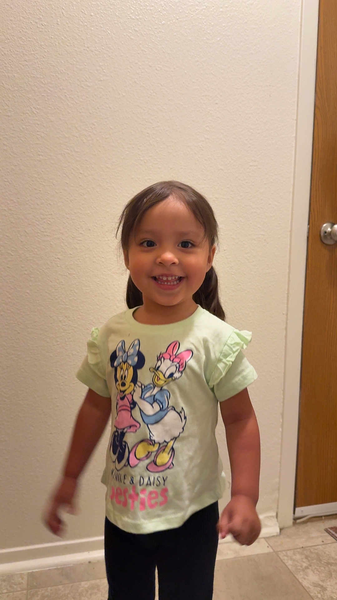 Liliana joined the competition — help win amazing prizes! child, smiling, girl, indoor, tshirt, cartoon_characters, minnie_mouse, daisy_duck, standing, happy, wall, door, floor, casual_clothing, portrait, person, young_child, playful, cute, home