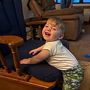 Landyn is registered to the contest to win money with this photo: toddler, child, smile, happy, indoor, rocking_chair, wood, cushion, white_shirt, camouflage_pants, living_room, carpet, furniture, person, baby_teeth, window, recliner, curtains, toy, playing