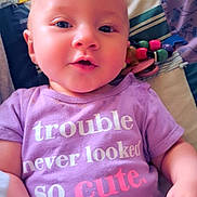 Journey is registered to the contest to win money with this photo: baby, blanket, child, closeup, clothing, colorful, cute, face, hand, happy, head, indoor, infant, person, portrait, purple_shirt, smiling, text_on_clothing, toy, young_child