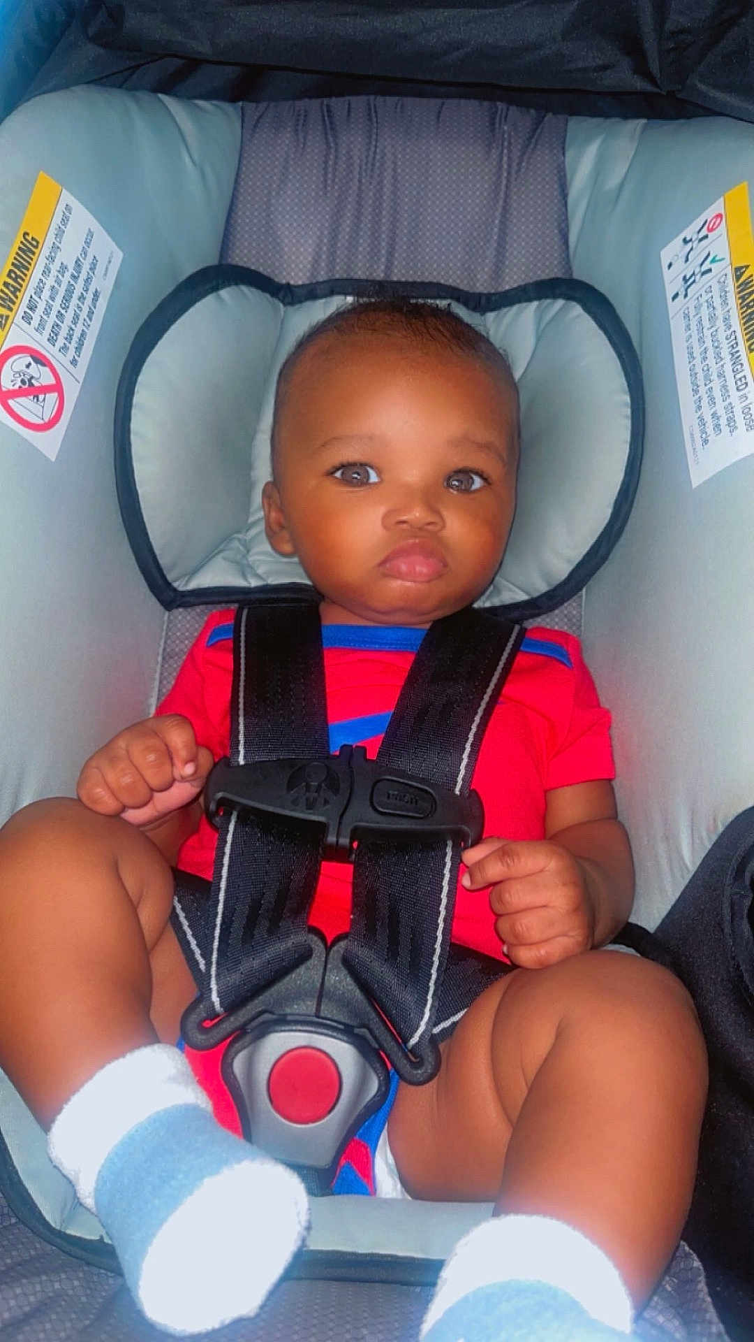 Prophet is registered to the contest to win money with this photo: baby, car_seat, child, infant, safety_harness, red_clothing, socks, seat_belt, cute, indoors, young_child, face, portrait, straps, head_support, hands, legs, expression, seat, person