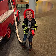Haydn joined the competition — help win amazing prizes! boots, boy, carpet, child, colorful_background, fire_hose, firefighter_costume, floor, fun, happy, indoor, jacket, kid, person, play_area, pretend_play, red_helmet, smiling, toy_fire_truck, young